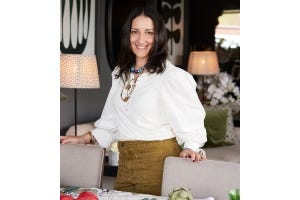 Kate Ferguson, wearing a white blouse, chunky necklaces and khaki-colored jeans, stands and smiles behind a table and dining chairs.