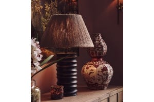 A black sculptural table lamp with a neutral shade sits next to a red patterned vase and a tortoiseshell candle holder 