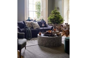 A velvet sofa piled high with cushions sits in a cosy sitting room, with a black-and-white coffee table sits and a Christmas tree decorated with warm fairy lights.