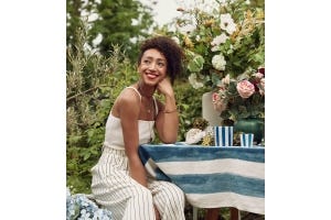 Hazel Gardiner is wearing a white top and white striped trousers, smiling and learning on her arm. Next to her is a floral-filled table.