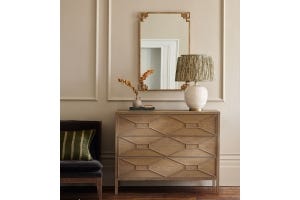 A rectangular mirror with metal gold frame up hanging on wall, in front of a chest drawers with a table lamp and a vase with flowers. 