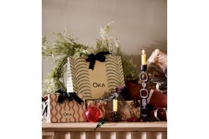 OKA gift bags are arranged on a table with foliage, candle holders, gifts and red patterned ornaments.