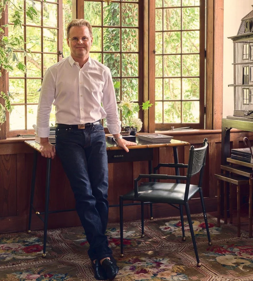 Adam Lippes wears a white shirt and blue jeans, and leans against a desk placed in front of a large window.