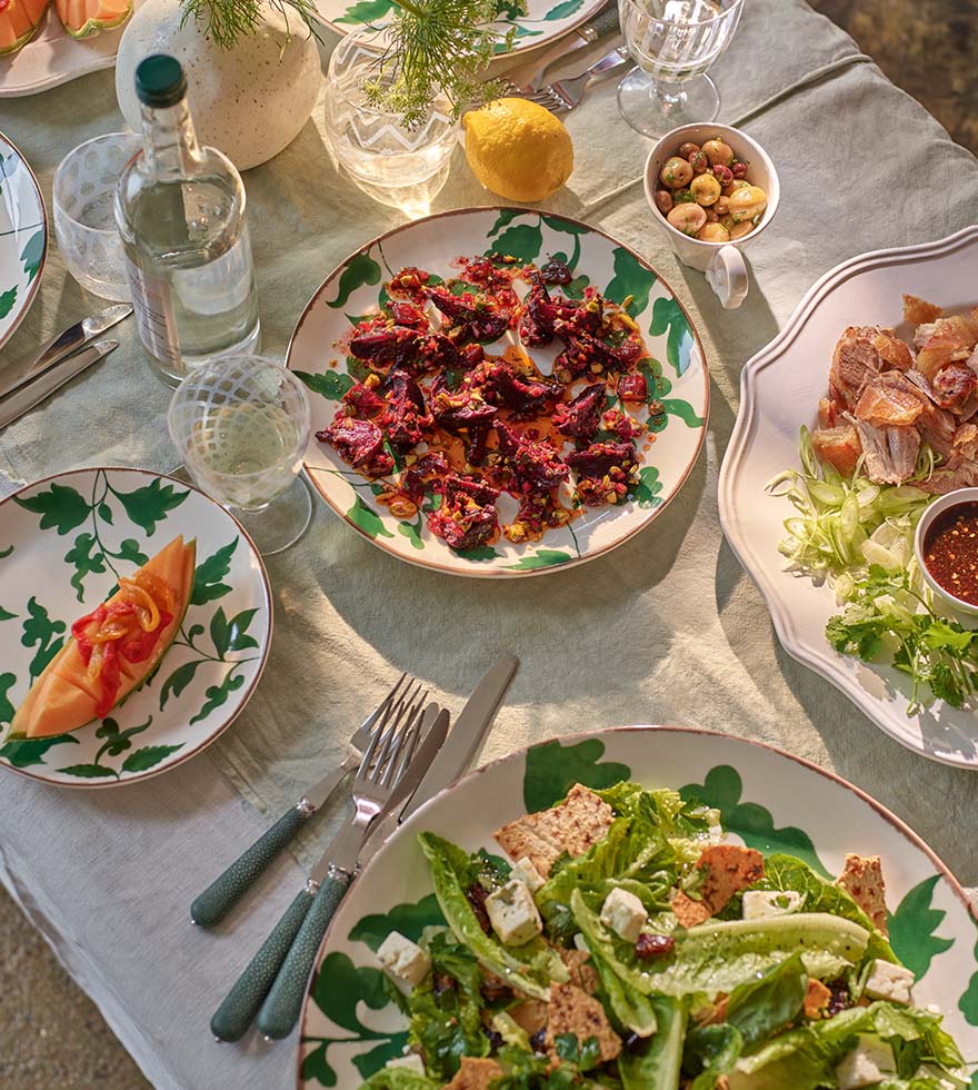 An array of healthy dishes including, melon, beetroot, and salad with toppings. Displayed on ivory print crockery on top of a white tablecloth. 