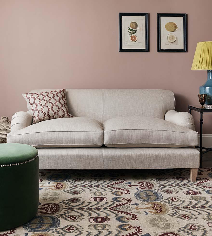 An off-white 3-seater sofa against a pale pink wall featuring fruit wall art. The sofa sits on top of a multicoloured patterned rug.