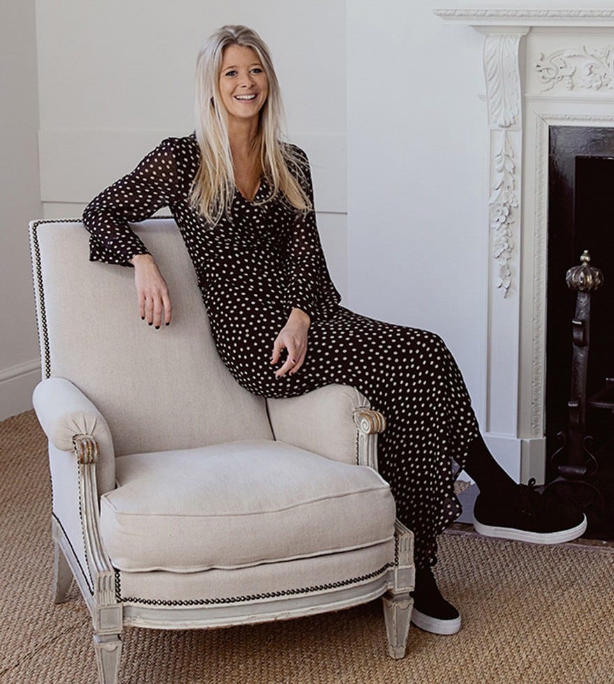 Camilla Clark is wearing a black and white polka dot dress, sitting on the arm of a natural colored accent chair with her legs crossed.