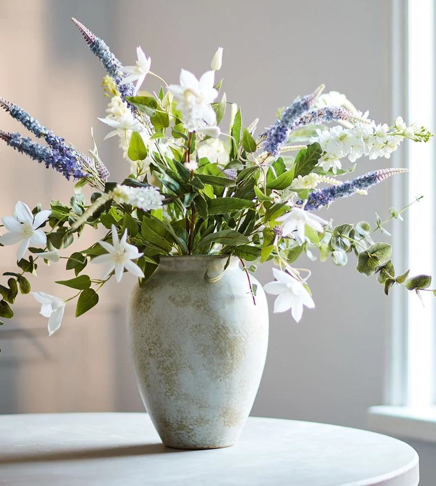 A vase with a large bouquet of faux blue and white flowers on top of a table.