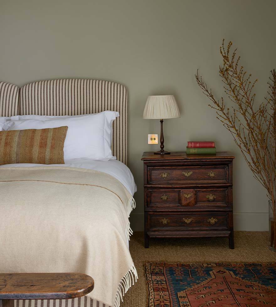 A double bed with a striped headboard with a cream throw and an orange stripe pillow. A dark wooden bedside table beside.