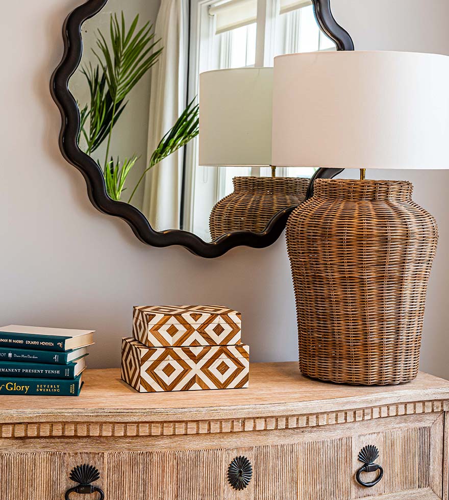 Decorative storage boxes, a stack of books and a rattan lamp on top of a console table. A black mirror with a scalloped edge hangs on the wall.