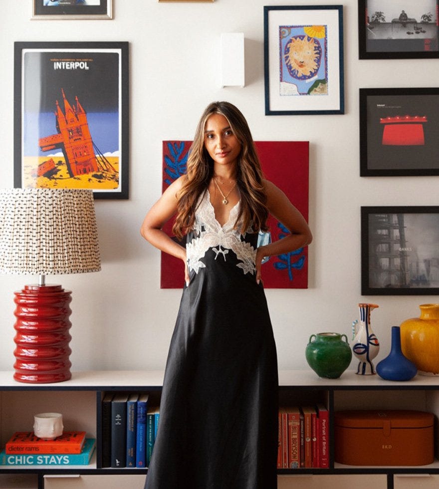 A woman in a black dress in front of a white wall with multiple colourful posters. A bright red lamp and multi-coloured ornaments on a sideboard behind.