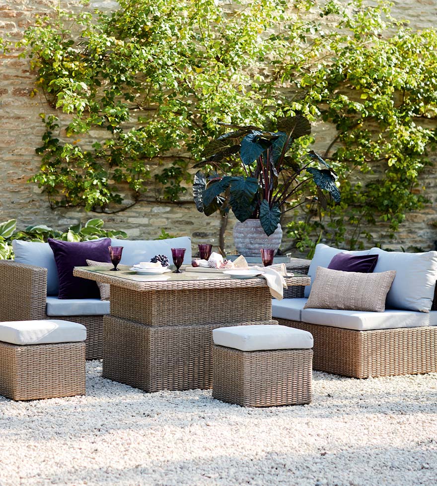 A lifestyle photograph set in a backyard with rattan outdoor furniture, laid with purple wine glasses and white tableware.