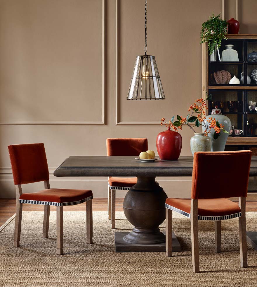 A lifestyle photograph of a dining room with a wooden dining table, orange chairs, vases, a low-hanging pendant lampshade and a wooden dresser in the background