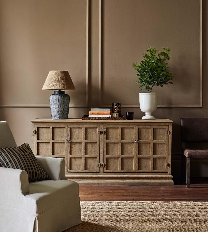 A corner of a sitting room with a large wooden console table, decorated with a blue lamp, white vase and books. In the foreground is a white armchair.