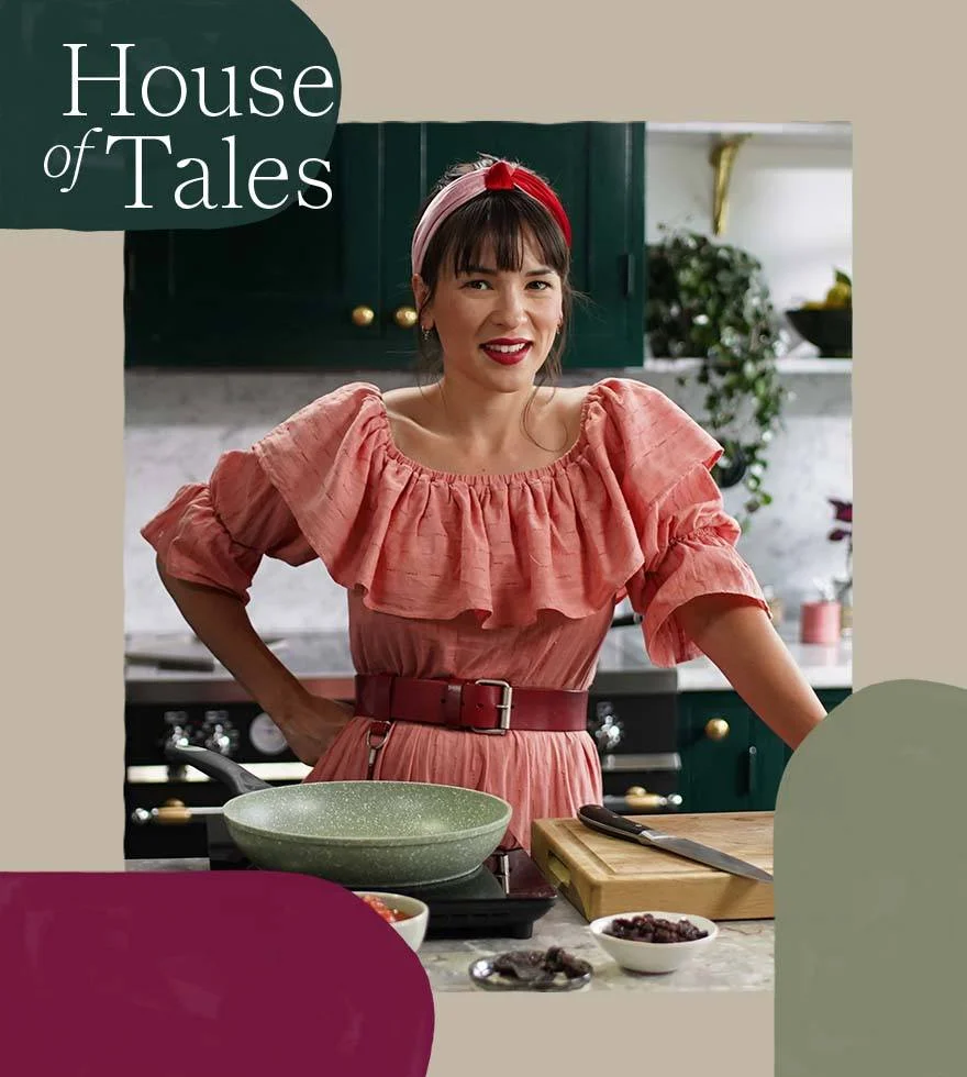 Rachel Khoo is wearing a pink dress with a red belt in her kitchen smiling with her hand on her hip.