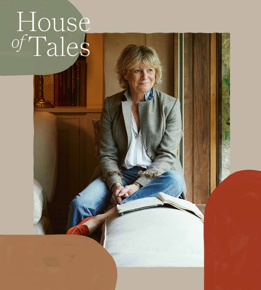 Sue Jones is sitting on a window bench, wearing a blazer, white shirt and blue jeans. She is looking out of the window, and a book is open in front of her. 