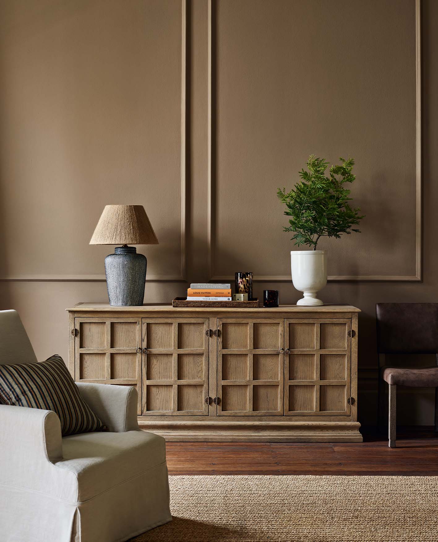 A corner of a sitting room with a large wooden console table, decorated with a blue lamp, white vase and books. In the foreground is a white armchair.