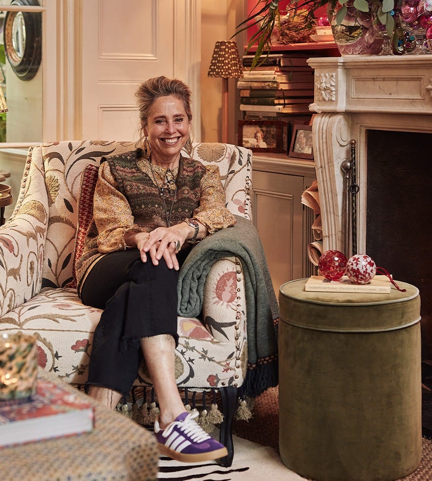 Butter Wakefield is smiling, wearing a Fair Isle-style sweater vest and sitting in an embroidered armchair, next to a green velvet ottoman.