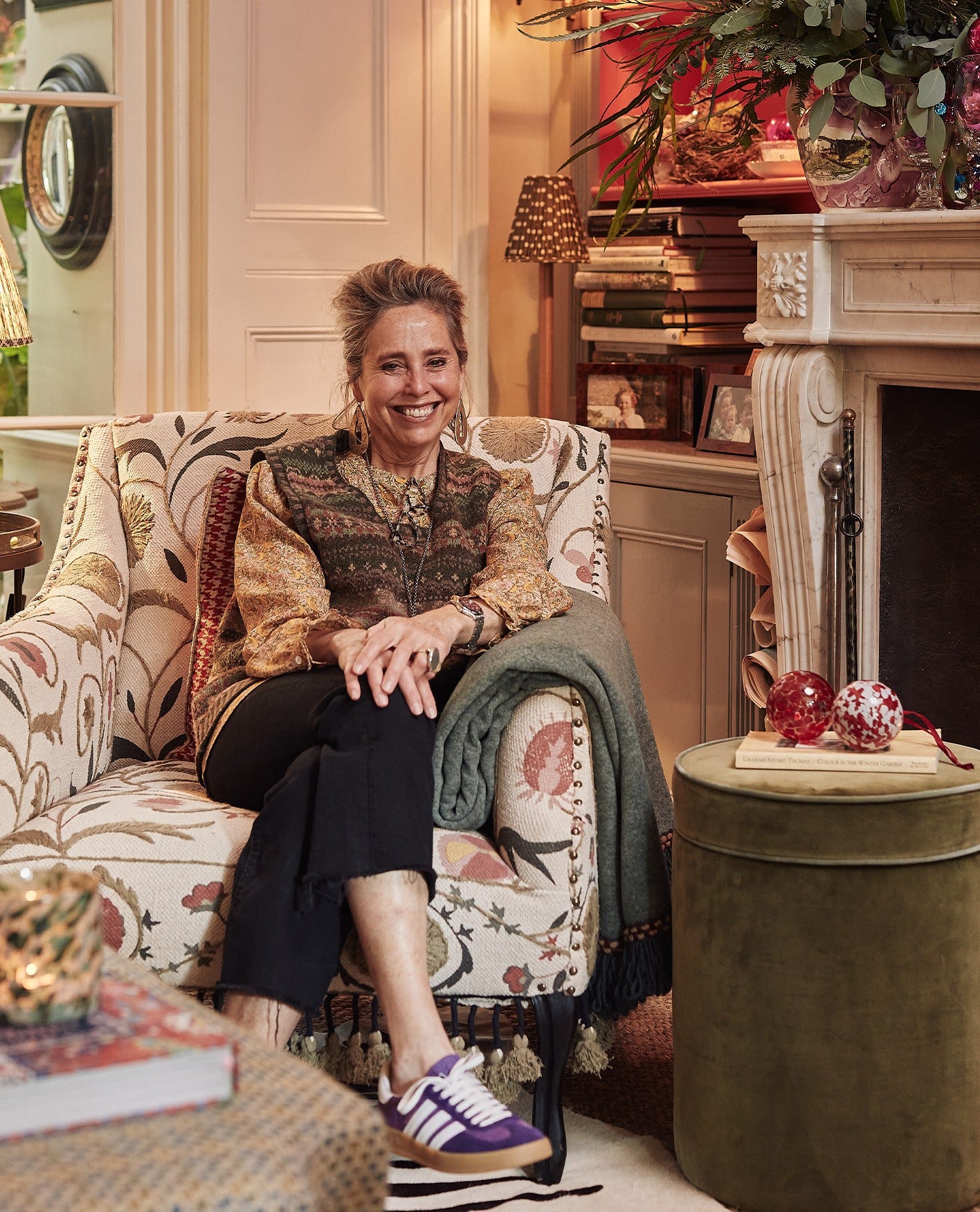 Butter Wakefield is smiling, wearing a Fair Isle-style sweater vest and sitting in an embroidered armchair, next to a green velvet ottoman.