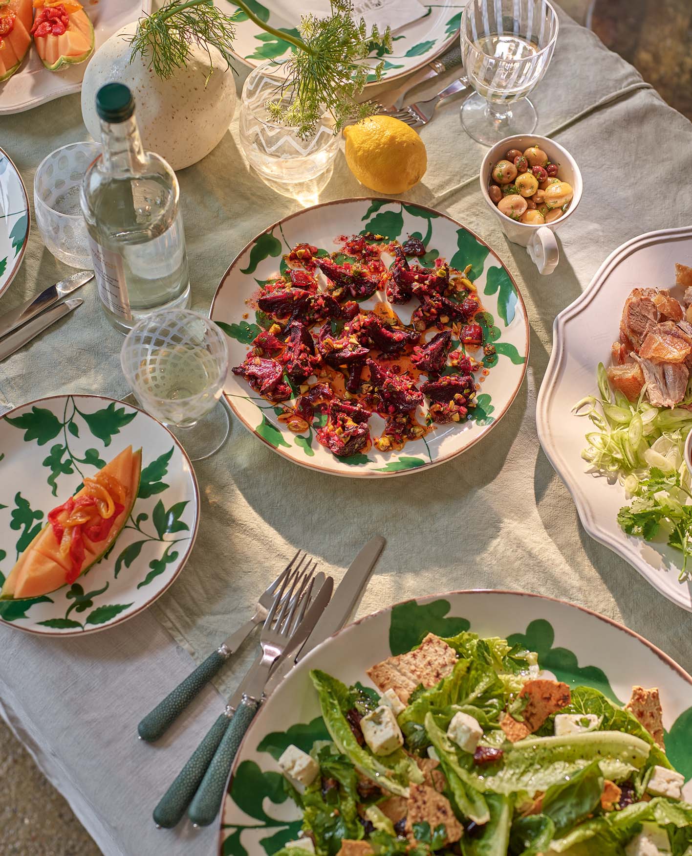 An array of healthy dishes including, melon, beetroot, and salad with toppings. Displayed on ivory print crockery on top of a white tablecloth. 