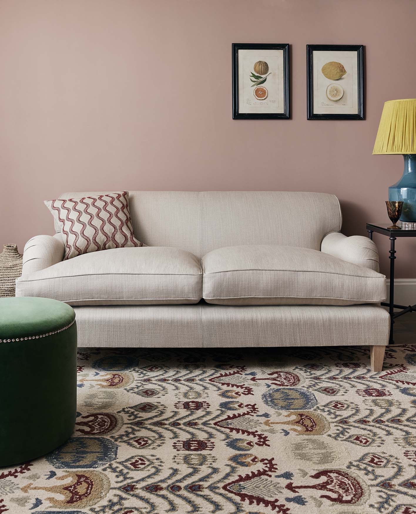 An off-white 3-seater sofa against a pale pink wall featuring fruit wall art. The sofa sits on top of a multicoloured patterned rug.