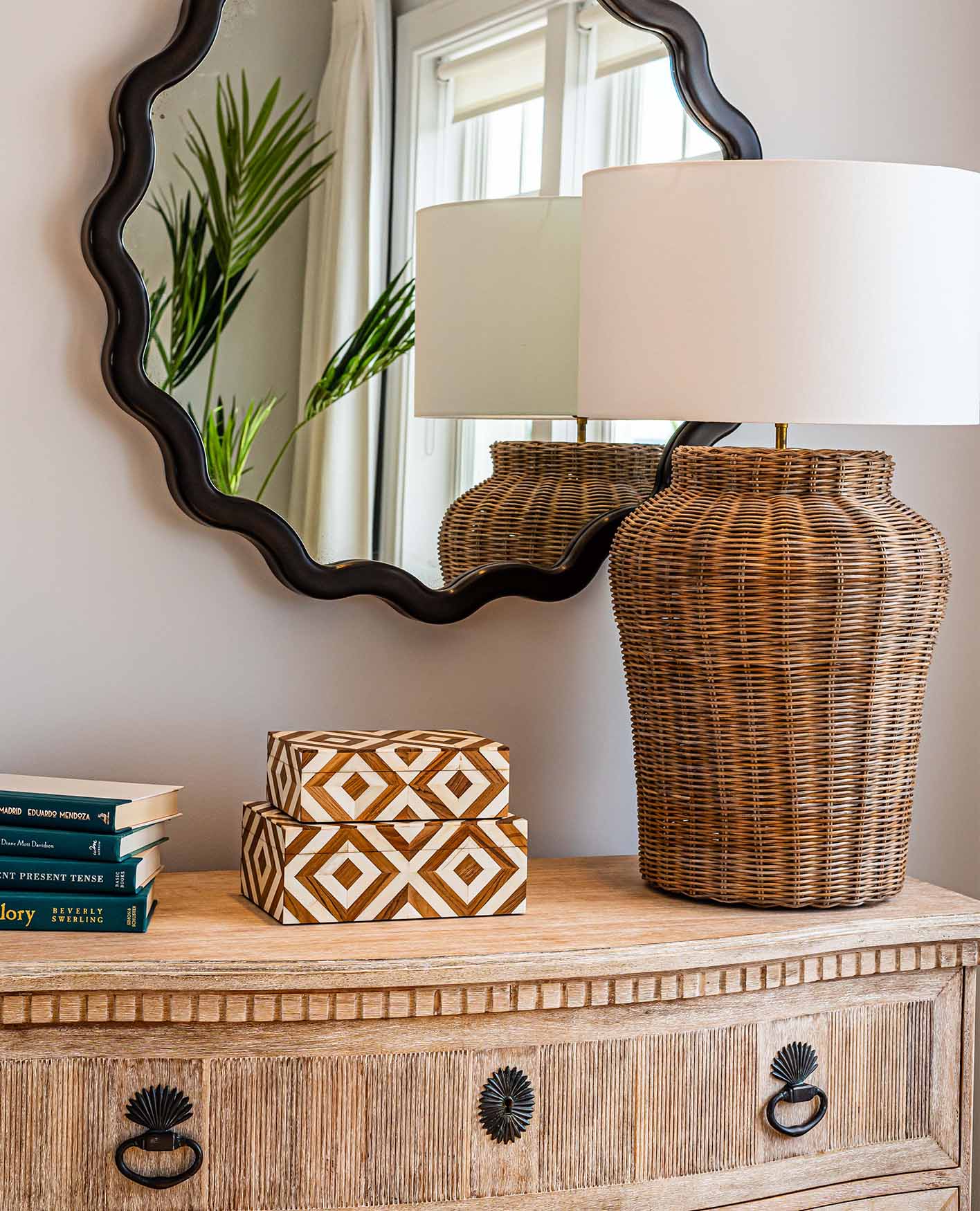 Decorative storage boxes, a stack of books and a rattan lamp on top of a console table. A black mirror with a scalloped edge hangs on the wall.