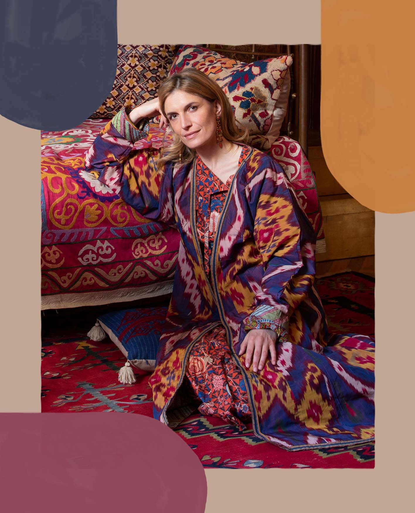 Martina Mondadori is sitting on the floor wearing a bright abstract patterned dress leaning with on a red patterned sofa.