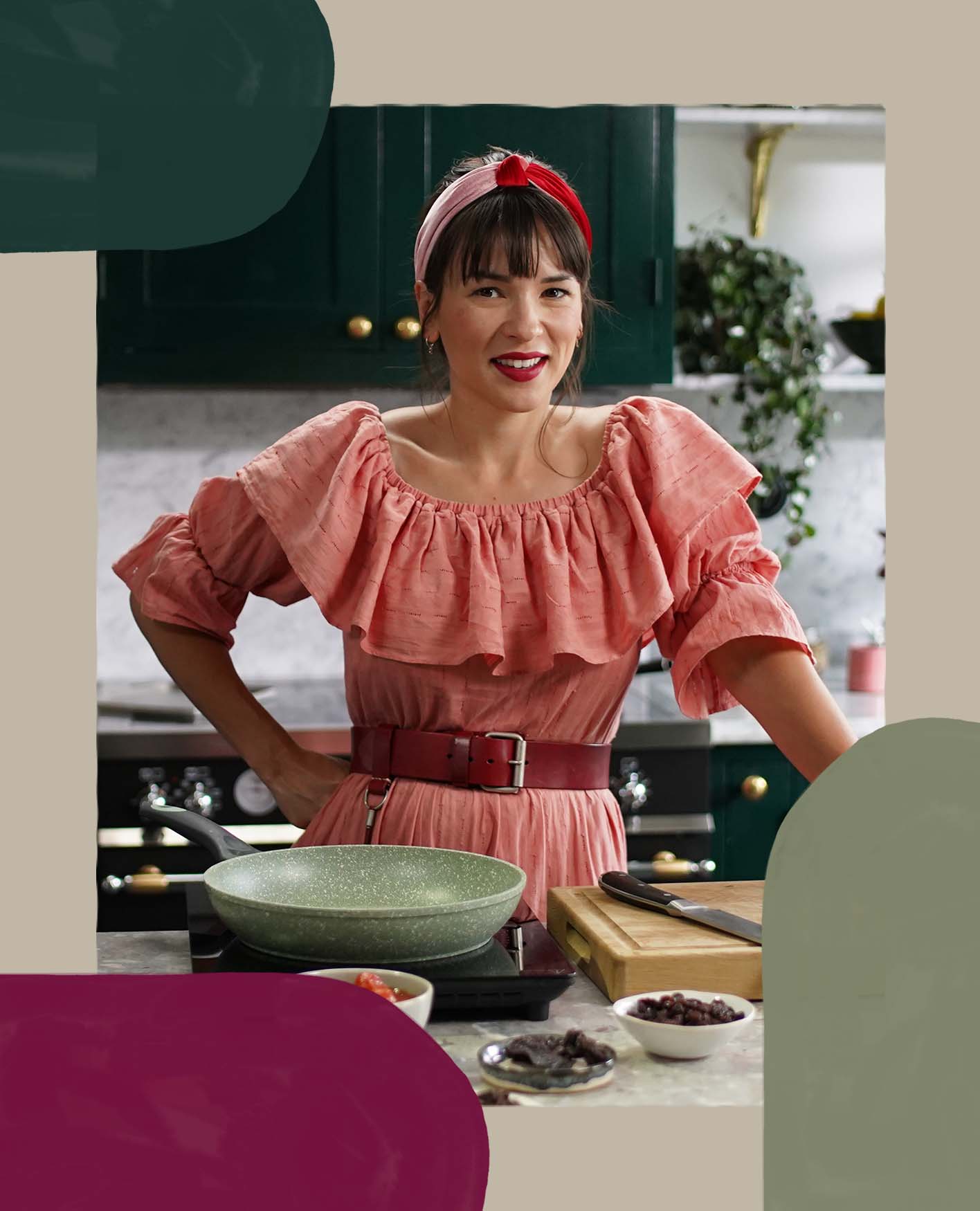 Rachel Khoo is wearing a pink dress with a red belt in her kitchen smiling with her hand on her hip.