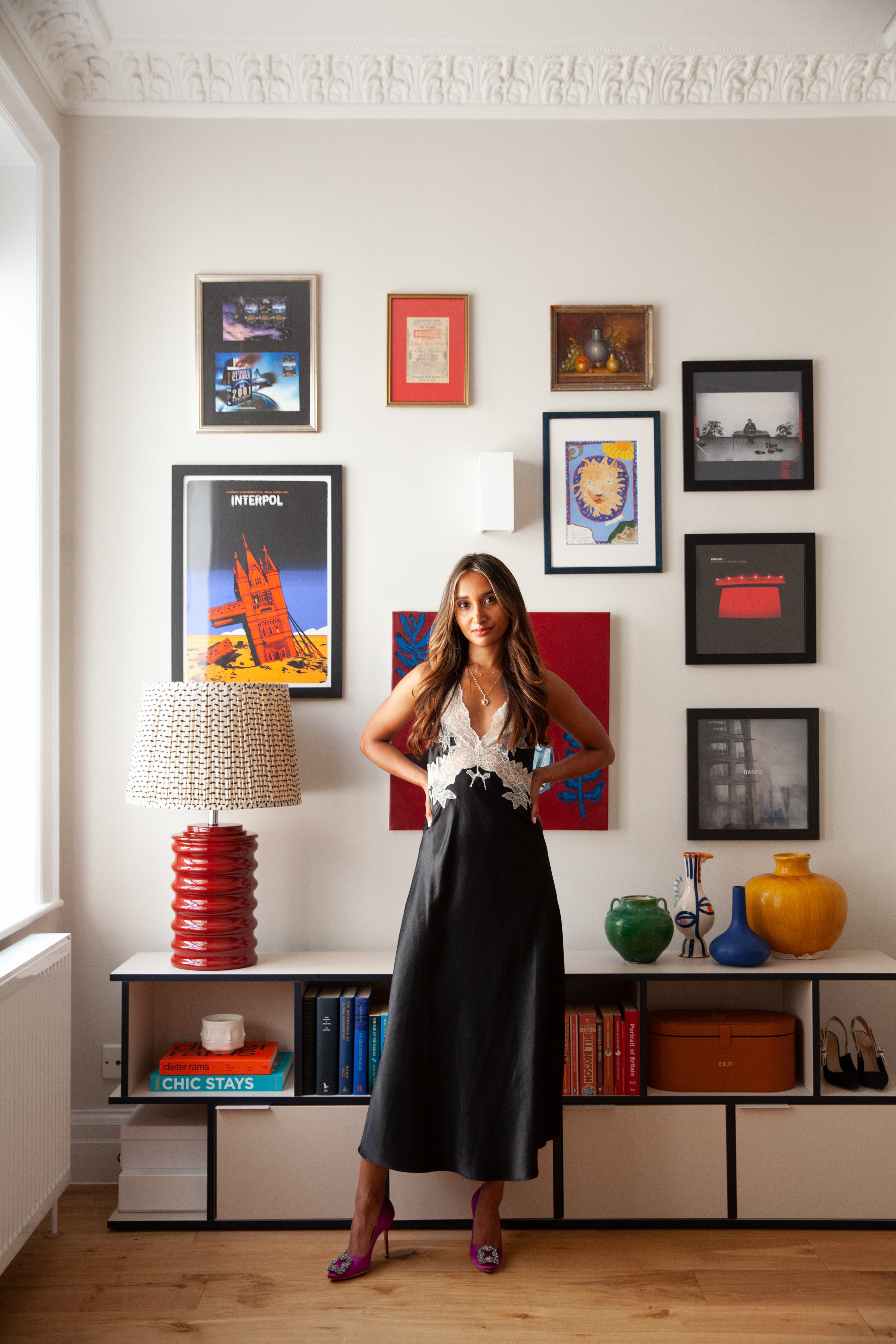 A woman in a black dress in front of a white wall with multiple colourful posters. A bright red lamp and multi-coloured ornaments on a sideboard behind.