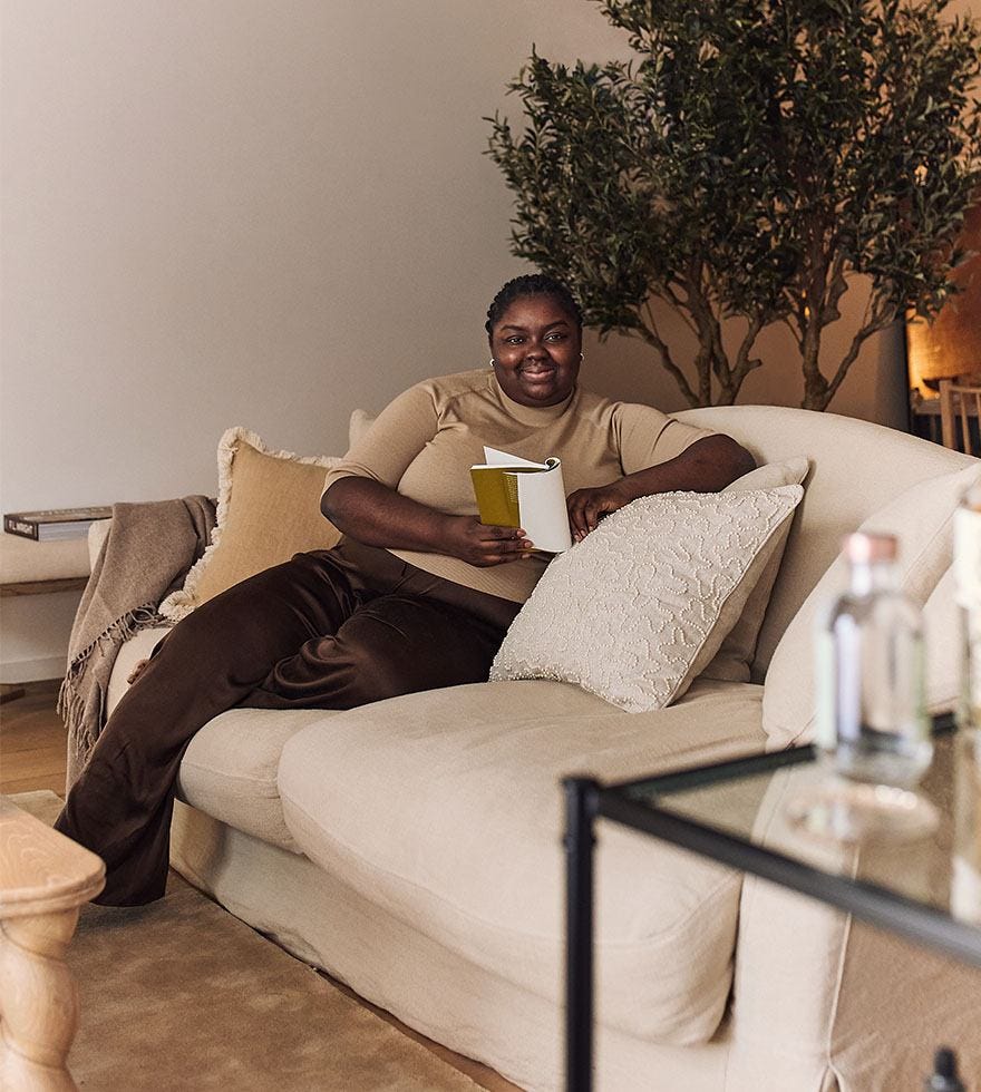 Absiola Omole is wearing brown trousers and a camel-colored top, reading a book and sitting on a linen sofa. A large faux tree is in the background.