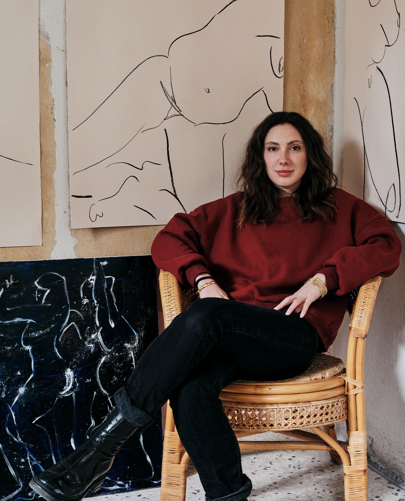 Alexandria Coe, wearing a red jumper and black trousers, sits in a wicker chair in front of her artworks.