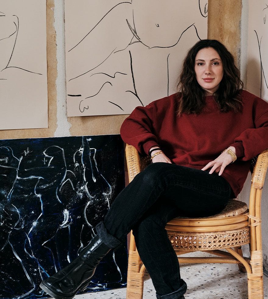 Alexandria Coe, wearing a red jumper and black trousers, sits in a wicker chair in front of her artworks.