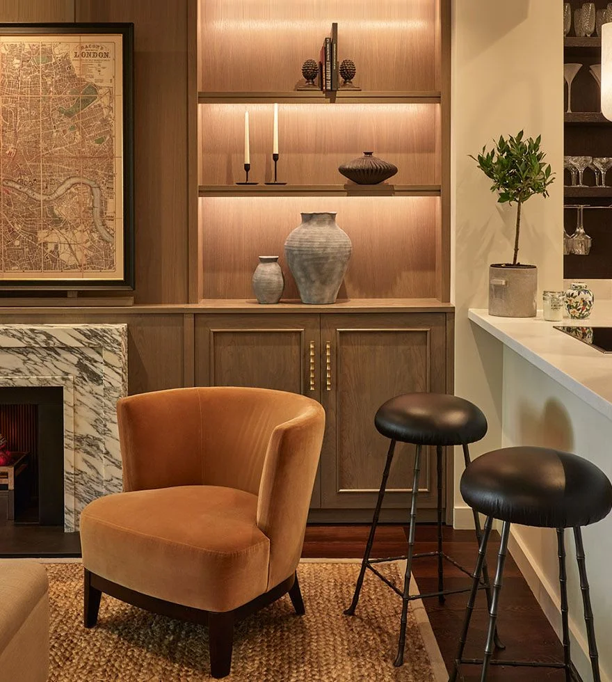 A cosy corner featuring an orange velvet armchair, a bookcase with integrated lighting and a kitchen counter with bar stools.