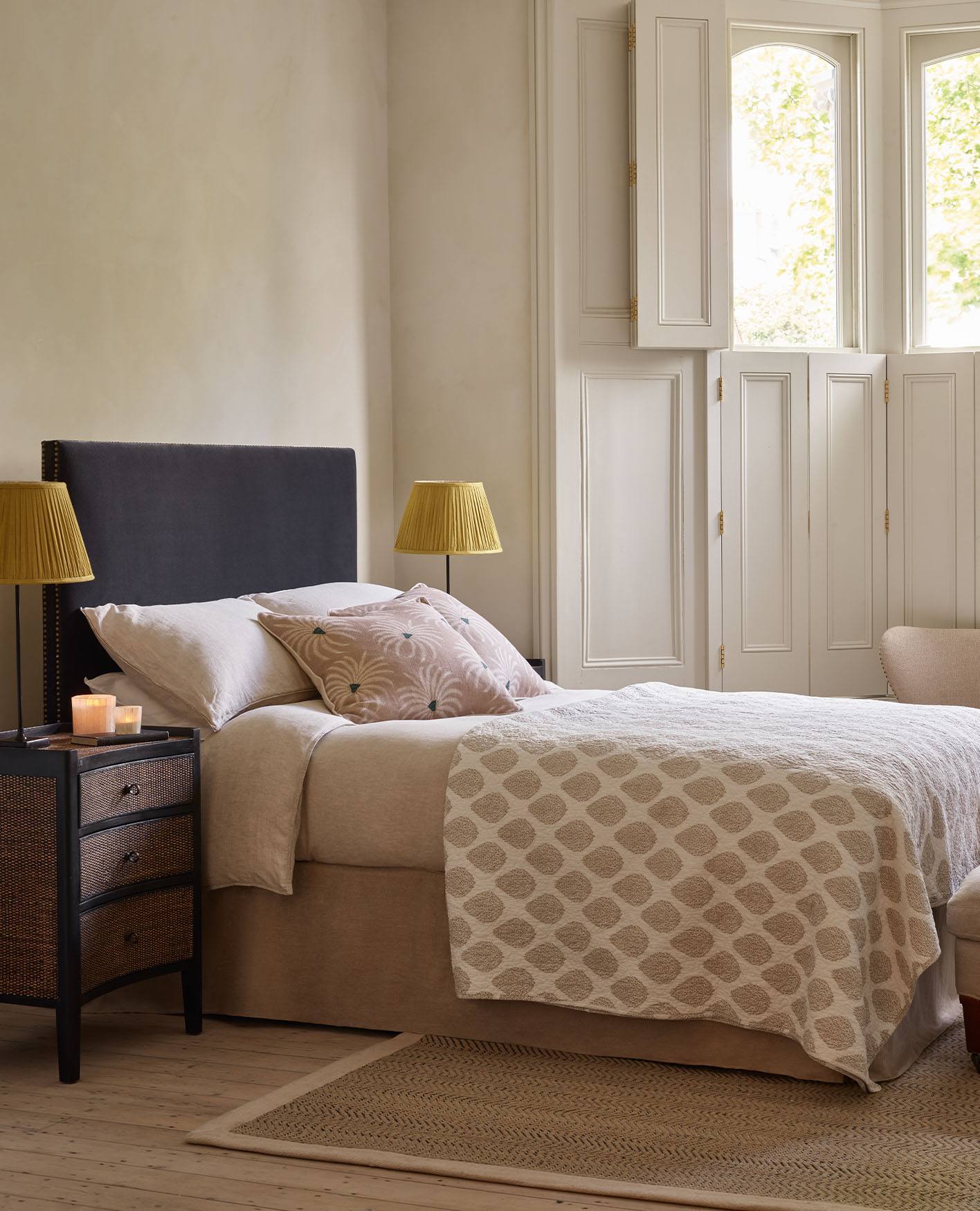 A cosy bedroom setting, featuring a navy blue headboard, patterned quilt and pillows and yellow pleated lampshades.