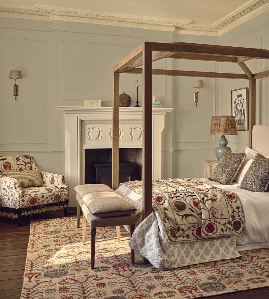 A softly coloured bedroom with a fourposter bed, floral-print red, olive green and cream armchair, bedspread and rug.