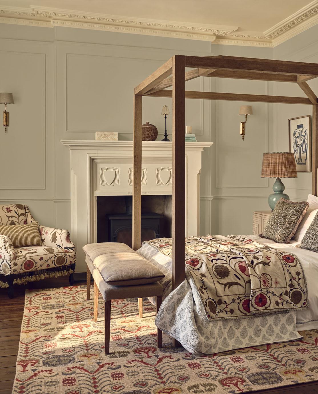 A softly coloured bedroom with a fourposter bed, floral-print red, olive green and cream armchair, bedspread and rug. 