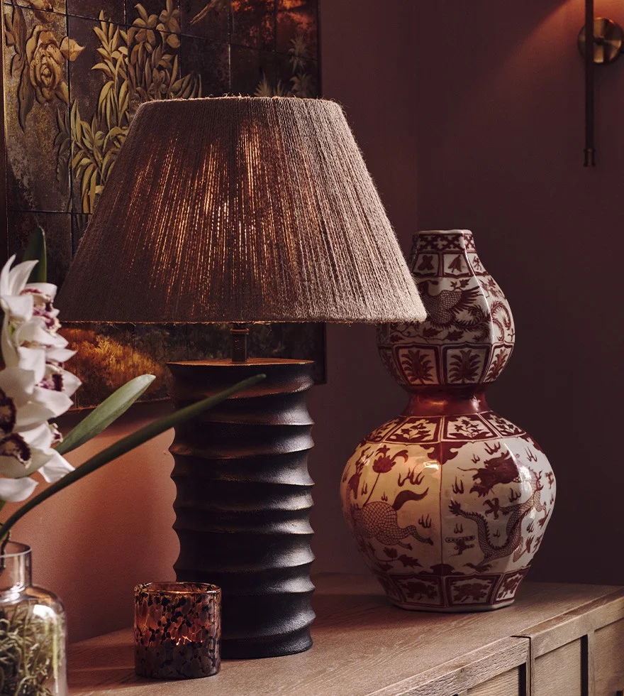 A black sculptural table lamp with a neutral shade sits next to a red patterned vase and a tortoiseshell candle holder 