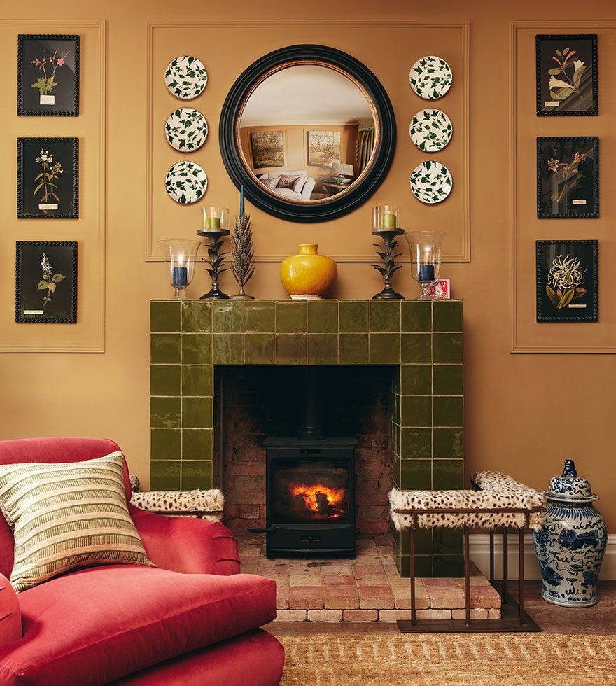 A vottage style living room featuring a red velvet armchair in front of a large green tiled fire place with a patterned fire fender. The mantle piece features decorative ornaments and the walls are decorated with a round mirror, patterned plates and art.