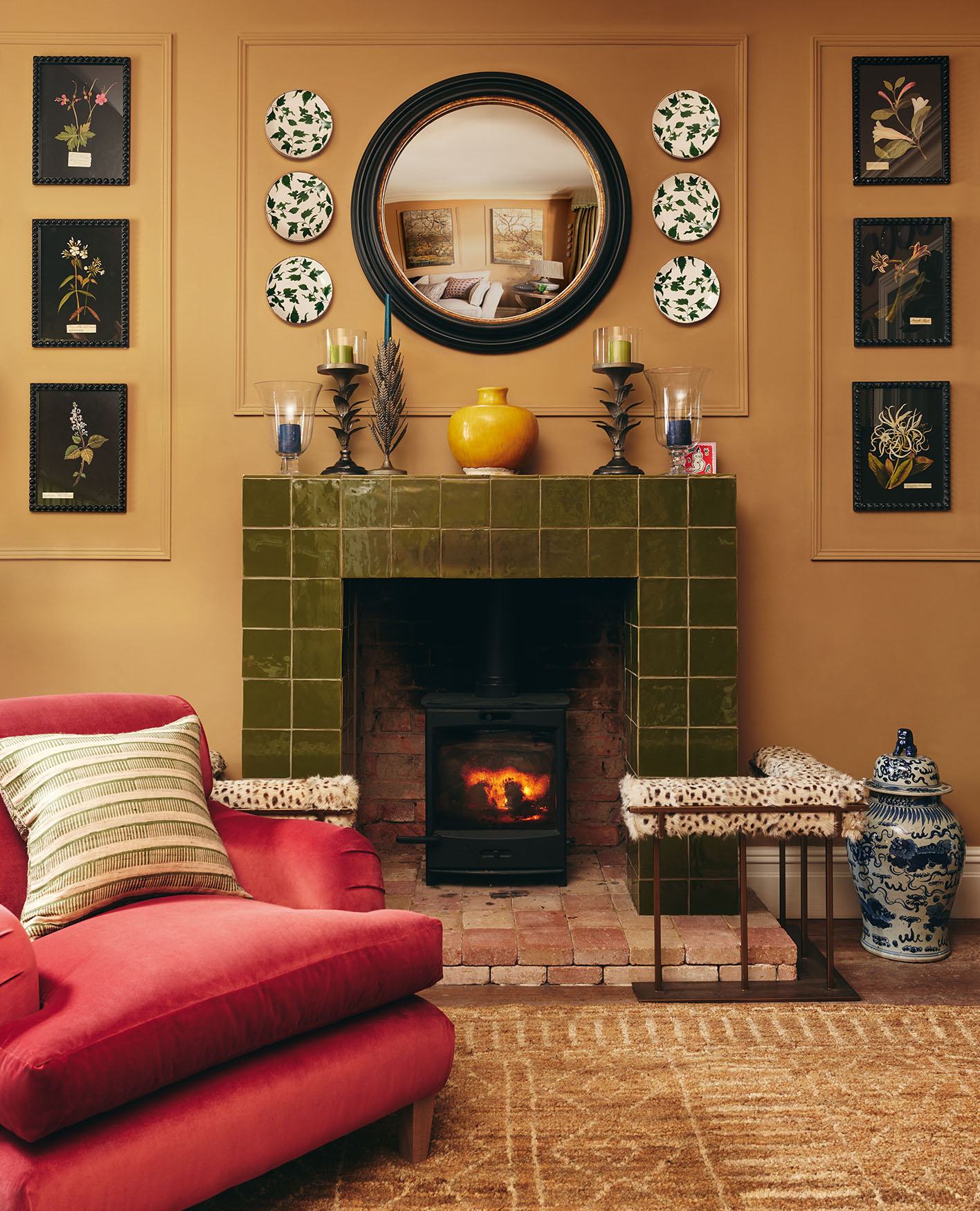 A vottage style living room featuring a red velvet armchair in front of a large green tiled fire place with a patterned fire fender. The mantle piece features decorative ornaments and the walls are decorated with a round mirror, patterned plates and art.