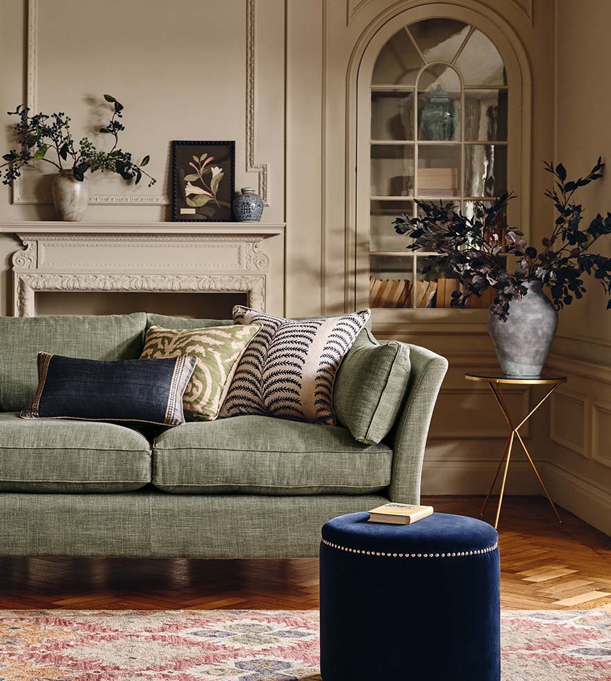 A cozy lounge with a glass bamboo coffee table with a red tray on top, a tan brown leather sofa and a patterned armchair, two lamps and window in the background