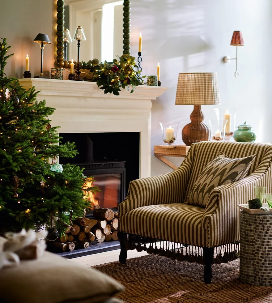 A green-and-white striped armchair sits in front of a roaring fire and a Christmas tree