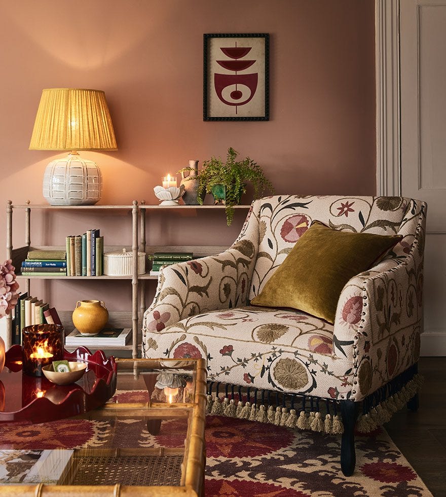 A corner of a cottage sitting room with a floral armchair, a shelf decorated with books, plants and a lamp and a framed art print hung on the wall.