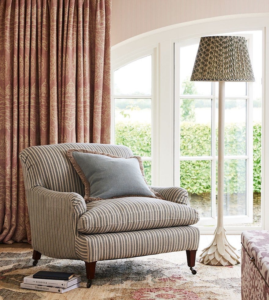 A corner of a living room, featuring a striped armchair next to a standing lamp, in front of a large window