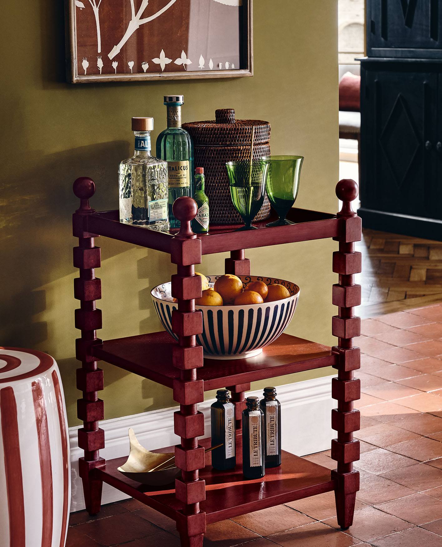 A red side table with cubed, bobbin-inspired legs holds an array of glassware and bottles of drink.