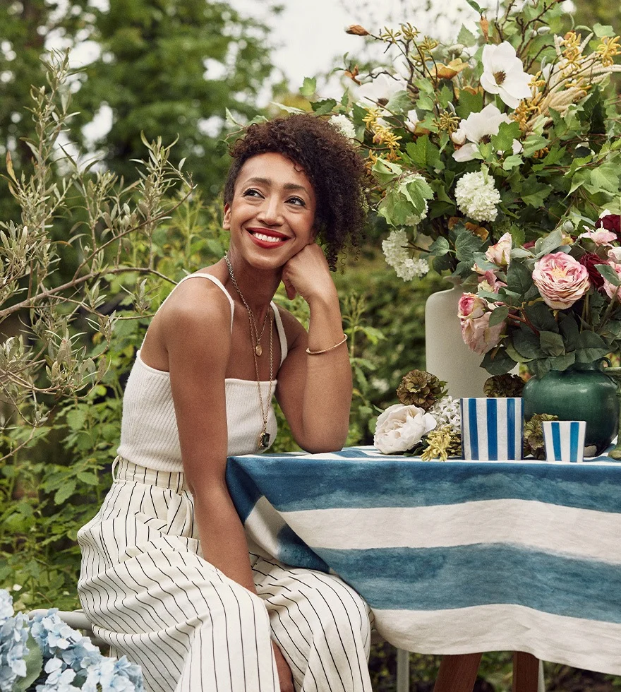 Hazel Gardiner is wearing a white top and white striped trousers, leaning on her arm and smiling, next to a floral-filled table.