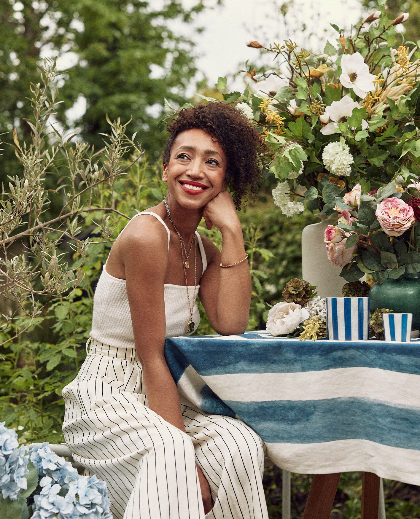 Hazel Gardiner is wearing a white top and white striped trousers, leaning on her arm and smiling, next to a floral-filled table.