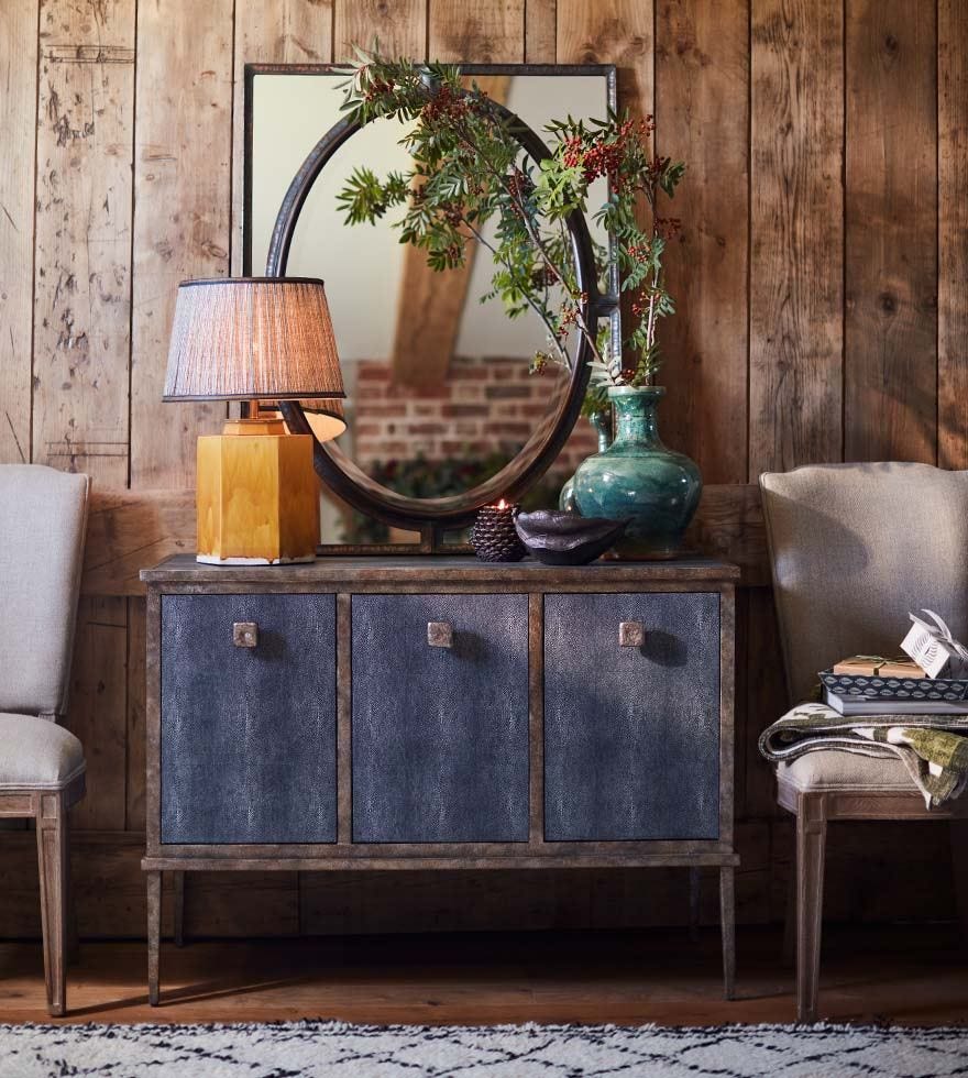 A sideboard cabinet featuring a yellow table lamp, a green vase, a large framed mirror and two dining chairs each side.