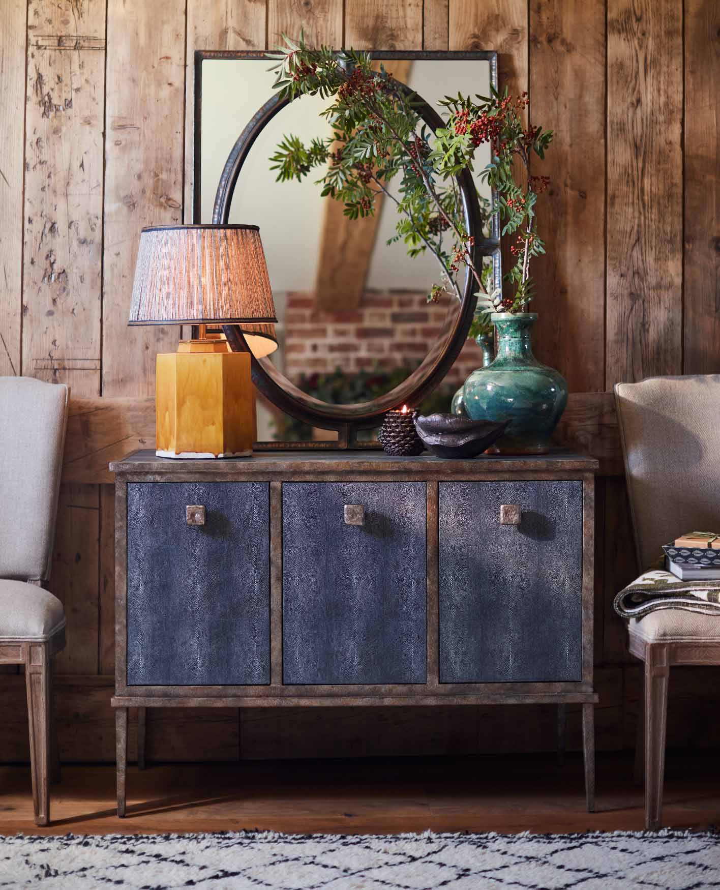 A sideboard cabinet featuring a yellow table lamp, a green vase, a large framed mirror and two dining chairs each side.