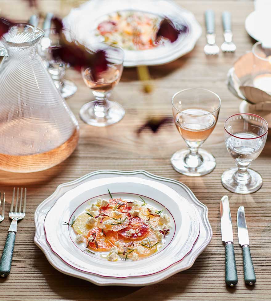 An arty photograph of two large clear wine glasses and two small clear wine glasses with a red rim, arranged on a wooden table set for dinner.