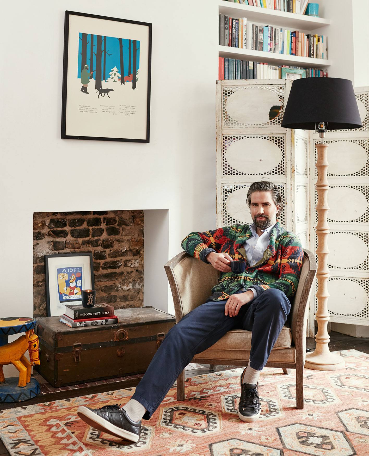 Jack Guinness sat in his sitting room on an armchair, next to a fireplace, with a floor lamp next to him and a screen and bookshelf behind.