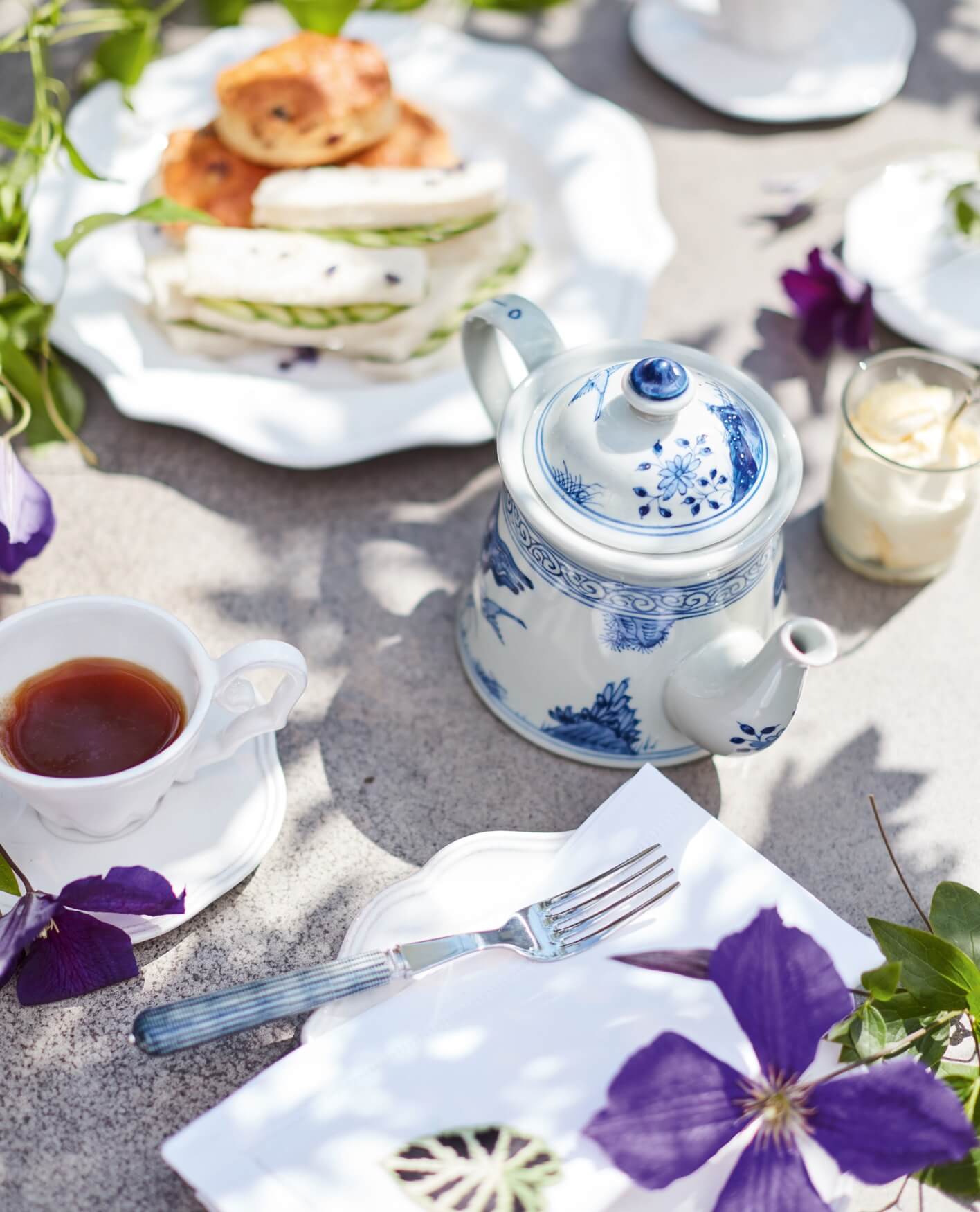 An elegant afternoon tea, with a blue-and-white teapot, and a white plate holding scones and cucumber sandwiches.